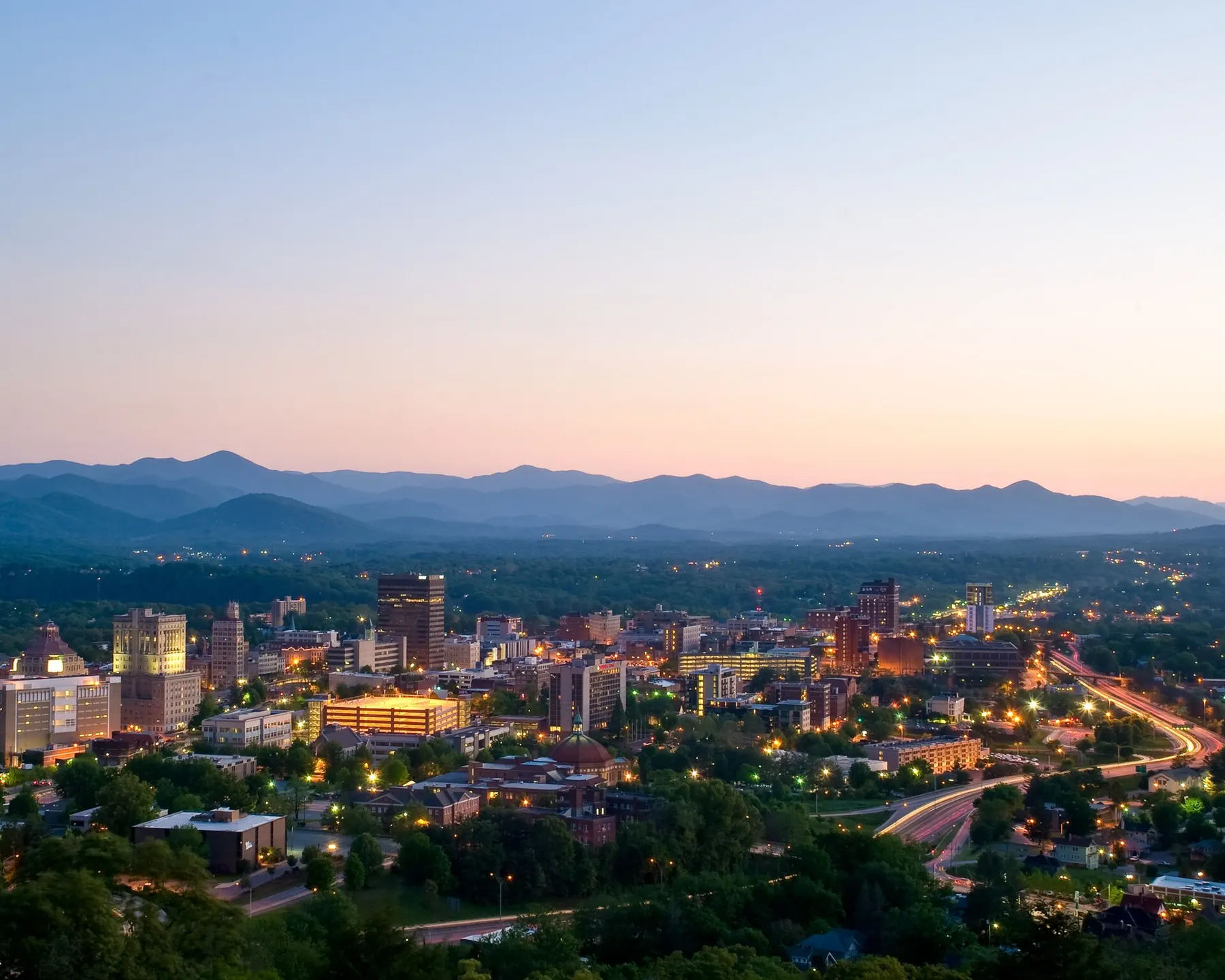 Downtown Asheville's buildings lit up at dusk, with layers of blue mountains behind them